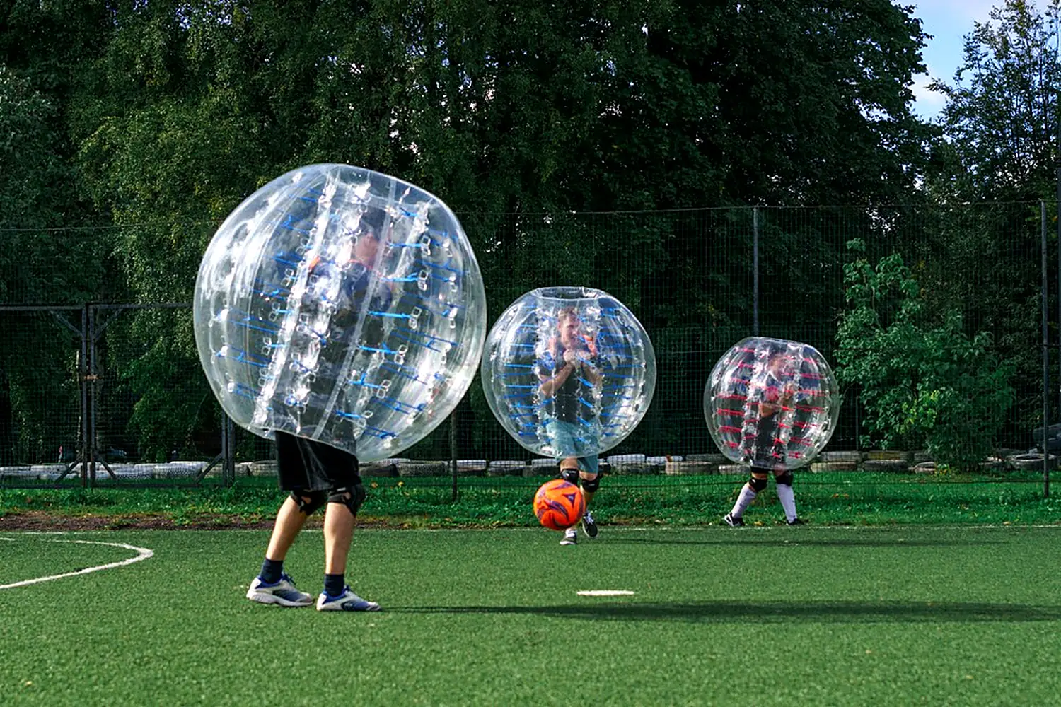 Three players wearing clear inflatable bubbles on a grass field during a bubble soccer game